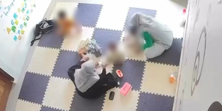 Overhead view of a room with checkered foam floor tiles; four blurred people sit on the floor in a circle, with a pink lunchbox and a bottle nearby.