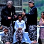 A girl seated in the center receives a ceremonial blessing from two men in traditional Balinese attire while others watch nearby.