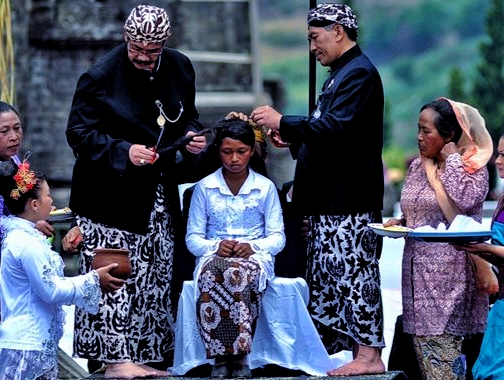 A girl seated in the center receives a ceremonial blessing from two men in traditional Balinese attire while others watch nearby.