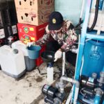 Maintenance worker in a patterned shirt and cap, wearing gloves, pours liquid into a blue bucket next to a white jug and water-pump equipment in a storage area.