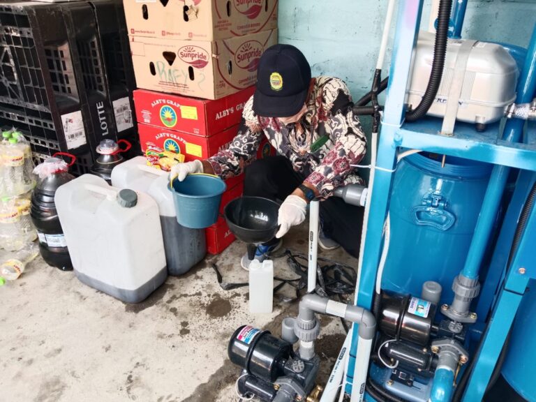 Maintenance worker in a patterned shirt and cap, wearing gloves, pours liquid into a blue bucket next to a white jug and water-pump equipment in a storage area.