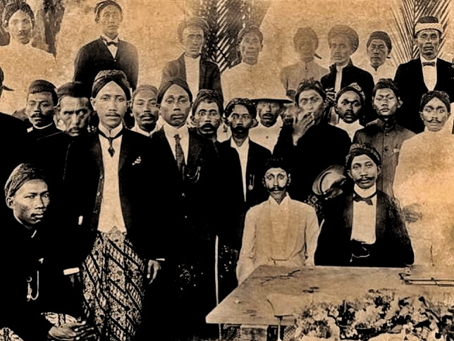 Historic group portrait of Black men and women in formal dress gathered around a coffin with a floral arrangement at a funeral.