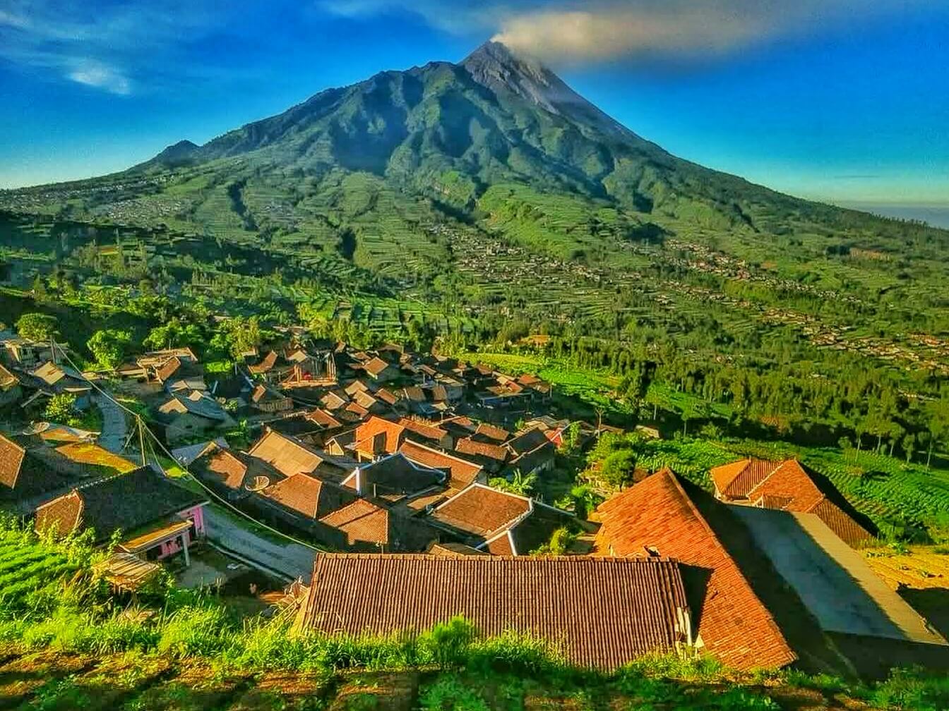 A hillside village with red-tiled roofs surrounded by green terraced fields under a tall mountain peak in clear blue sky.