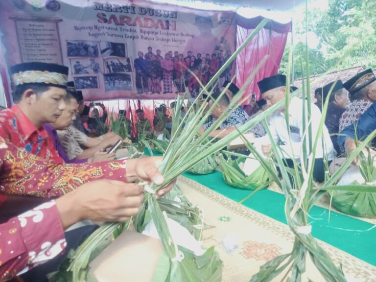 Group of Indonesian people in traditional attire weaving palm leaves at a long table during a cultural ceremony under a tent with a banner behind them.