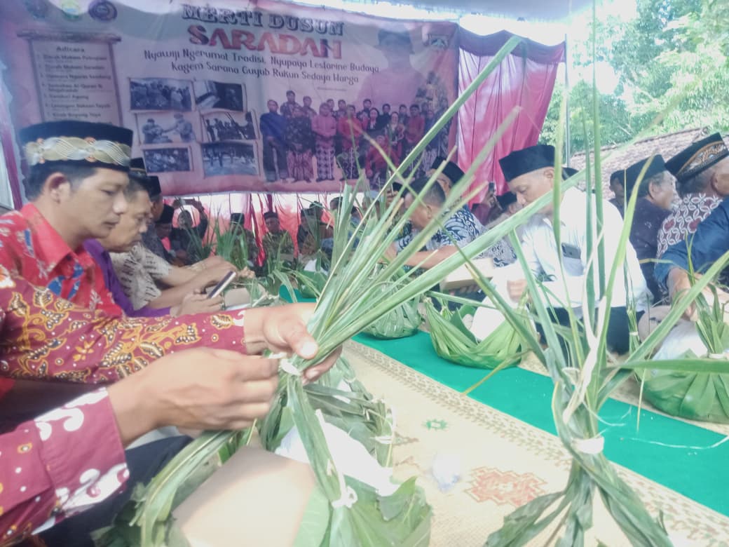 Group of Indonesian people in traditional attire weaving palm leaves at a long table during a cultural ceremony under a tent with a banner behind them.