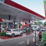 Gas station with multiple pumps and a white SUV refueling under a large canopy; fuel price board visible on the right.
