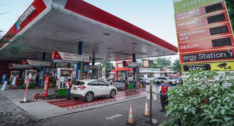 Gas station with multiple pumps and a white SUV refueling under a large canopy; fuel price board visible on the right.