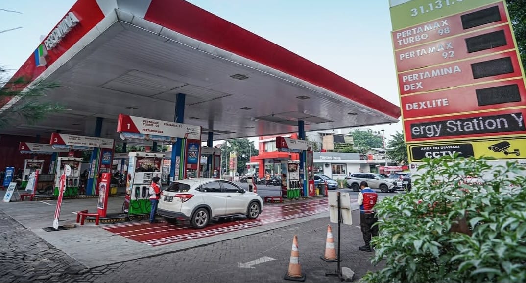 Gas station with multiple pumps and a white SUV refueling under a large canopy; fuel price board visible on the right.
