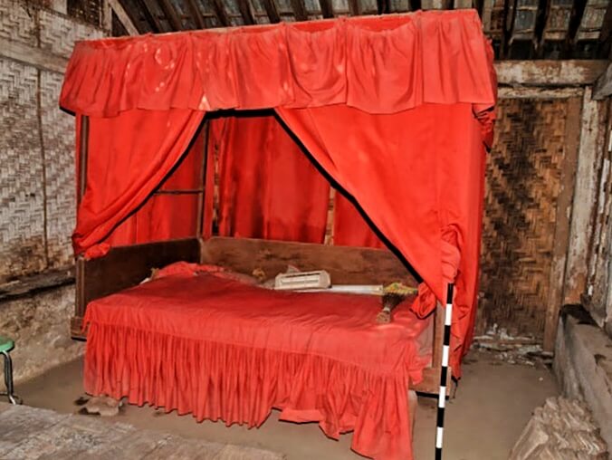 Red canopy bed with draped red curtains in a rustic room, a low wooden frame and ruffled bed skirt visible.