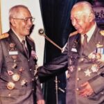 Two senior military officers wearing dark green dress uniforms with medals and gold shoulder boards, smiling as they shake hands.