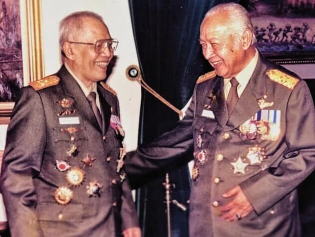 Two senior military officers wearing dark green dress uniforms with medals and gold shoulder boards, smiling as they shake hands.