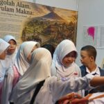 Group of Indonesian female students in white hijab uniforms gathered around a museum display featuring a volcano painting and informational panels.