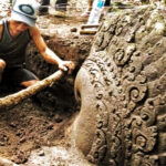 Person kneeling in mud, digging around a large ornate carved stone wheel.