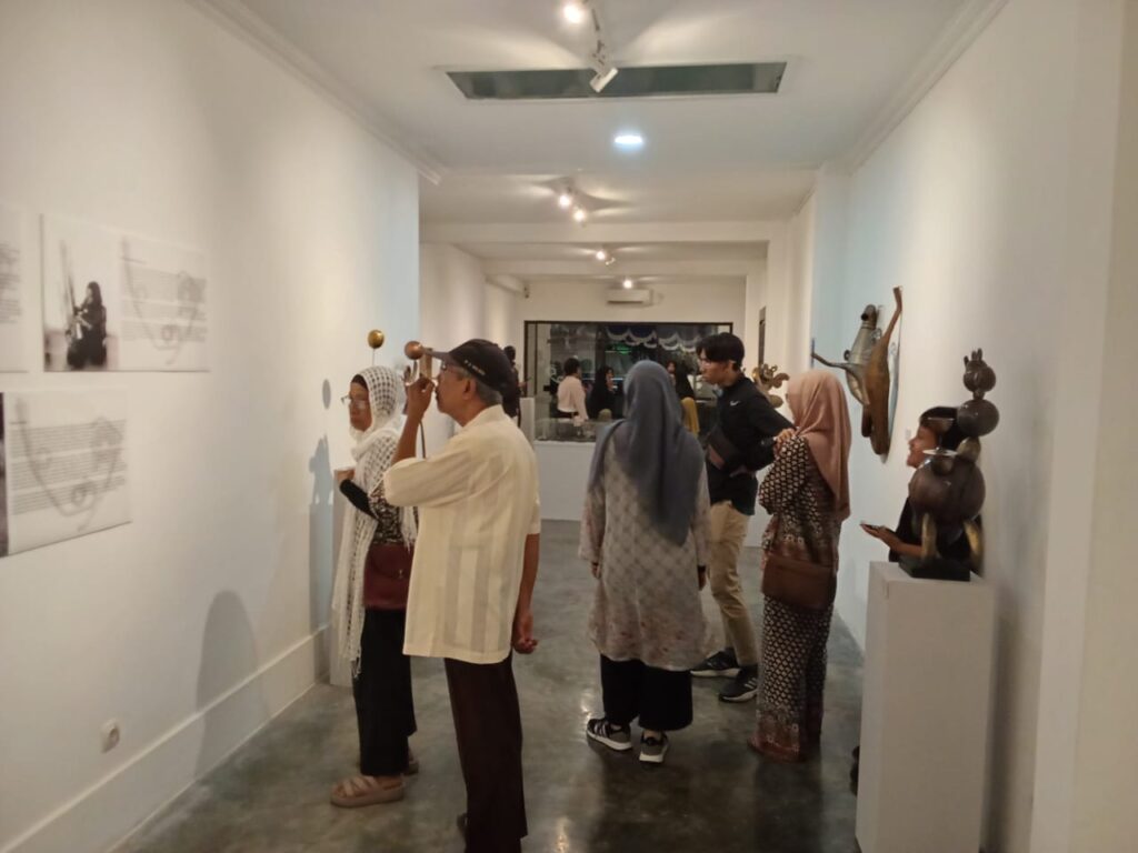 Visitors in a bright gallery observing wall-mounted artwork; white walls, sculptures on a pedestal visible to the right.