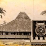 Historic building with a tall thatched roof and a large shield emblem mounted on a pedestal in front.