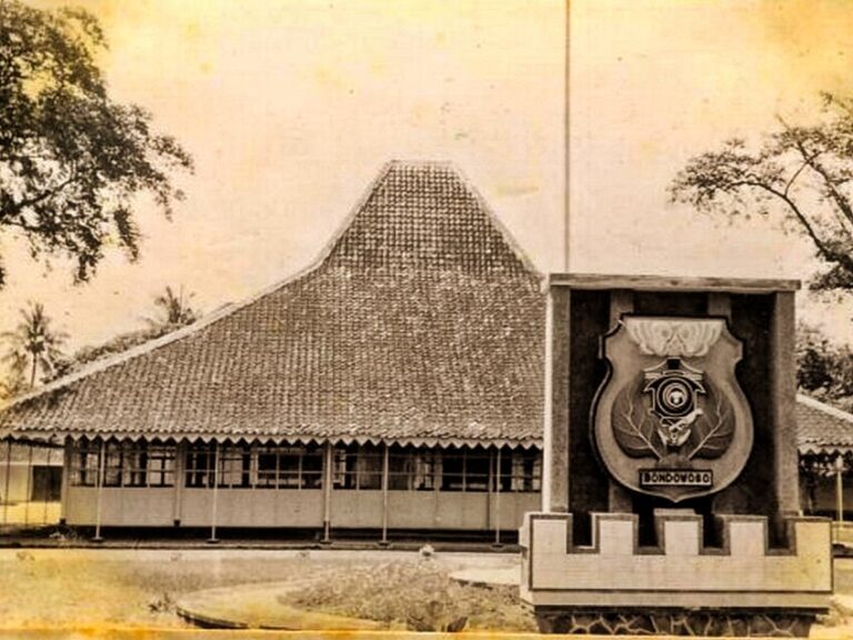 Historic building with a tall thatched roof and a large shield emblem mounted on a pedestal in front.