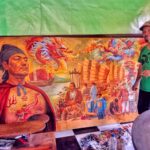 Man in a green shirt and cap stands beside a large, colorful mural featuring a dragon, ships, and people by the waterfront.