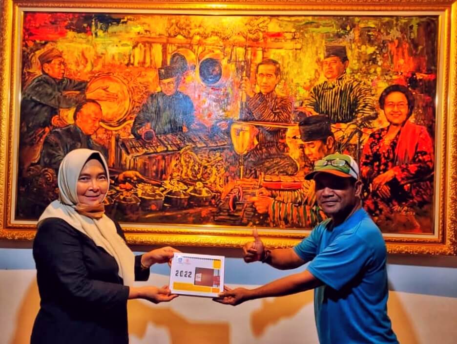 Woman in a hijab hands a 2022 calendar to a man in a blue polo, both smiling in front of a large framed painting with warm colors.