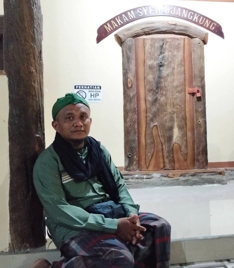 Two people sit on a white floor beside massive wooden pillars inside a hall, with a sign reading Makam Syed Djangkung above the door.