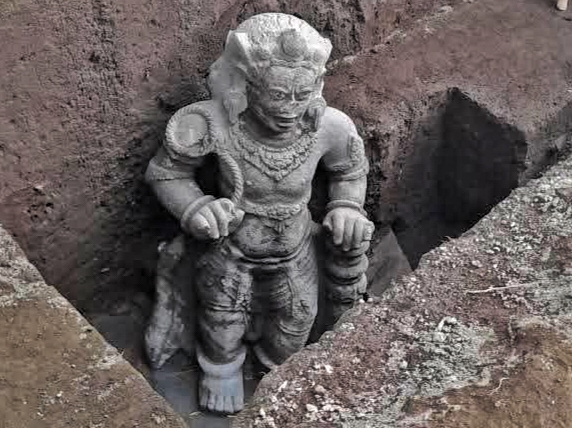 Stone carved statue of a muscular mythic figure standing in a rectangular dirt pit, with jewelry and outstretched arms, weathered and partially buried in earth.