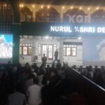 Nighttime outdoor community event in front of Nurul 'Ashri Deresan, with a large audience seated and two projection screens displaying blue imagery on either side of a lit stage.