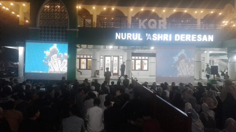 Nighttime outdoor community event in front of Nurul 'Ashri Deresan, with a large audience seated and two projection screens displaying blue imagery on either side of a lit stage.
