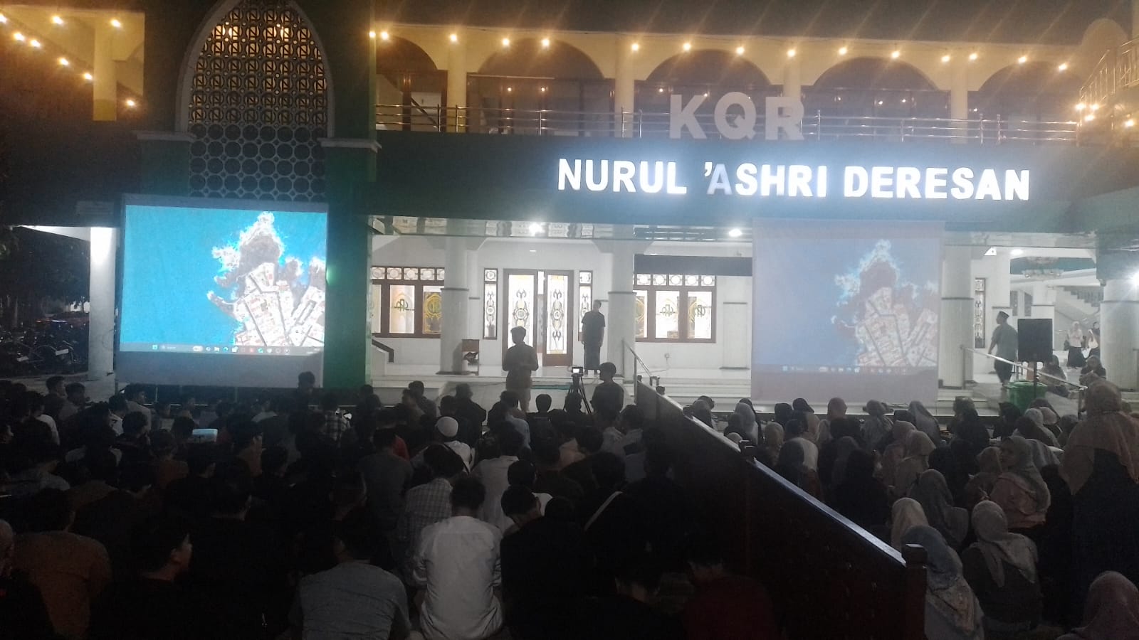 Nighttime outdoor community event in front of Nurul 'Ashri Deresan, with a large audience seated and two projection screens displaying blue imagery on either side of a lit stage.