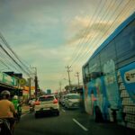 Busy urban street scene with cars, motorcycles, and a blue bus driving past storefronts under a tangle of power lines at daybreak.