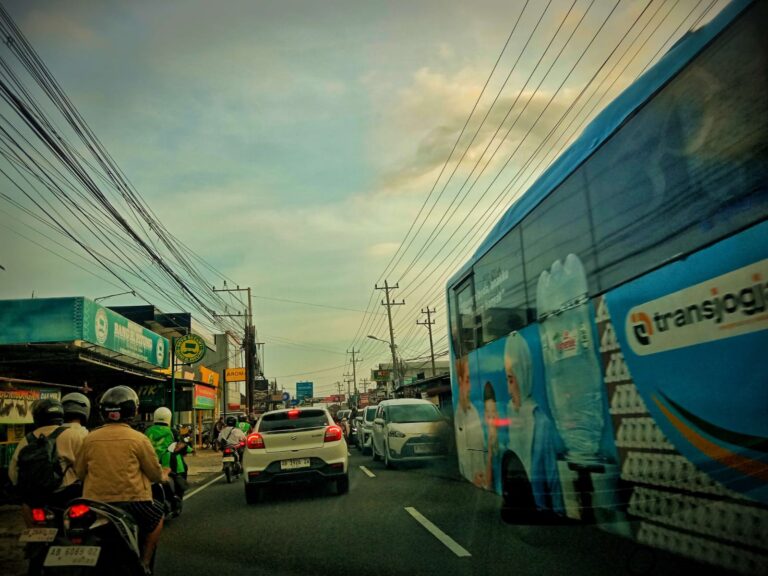 Busy urban street scene with cars, motorcycles, and a blue bus driving past storefronts under a tangle of power lines at daybreak.