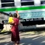 Woman in a red dress lifts a baby wrapped in yellow toward a passing green-and-white train as a man on a scooter looks on nearby.