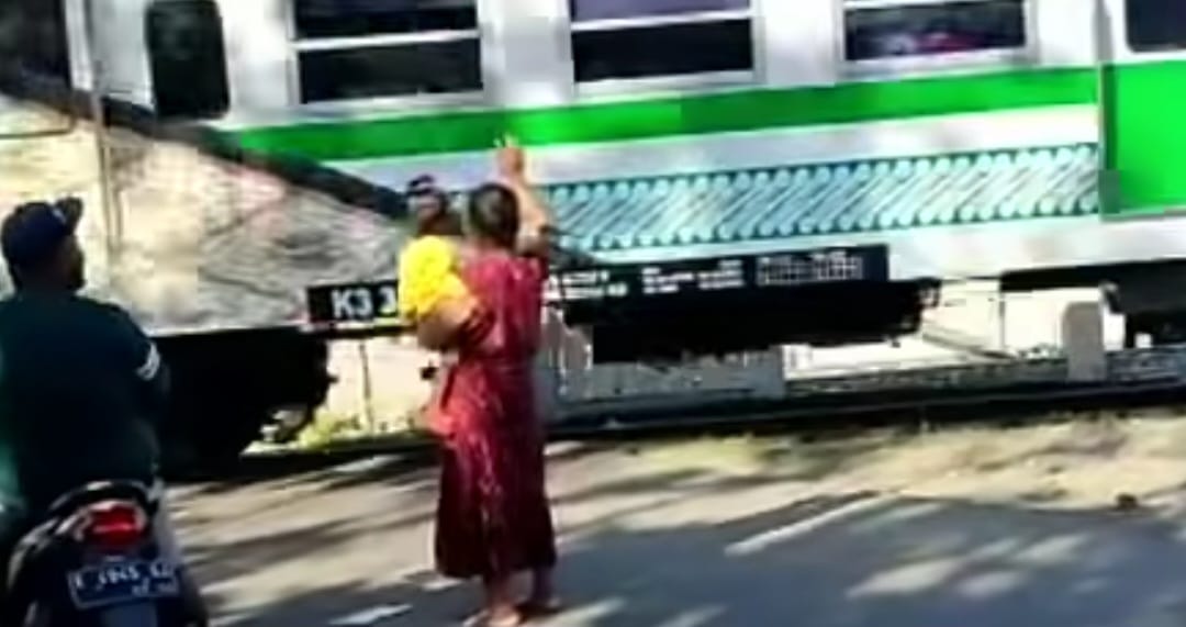 Woman in a red dress lifts a baby wrapped in yellow toward a passing green-and-white train as a man on a scooter looks on nearby.