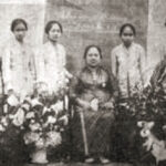 Old black-and-white group portrait: a seated elderly woman with a walking cane, surrounded by six children, with flowers and a small dog in front.