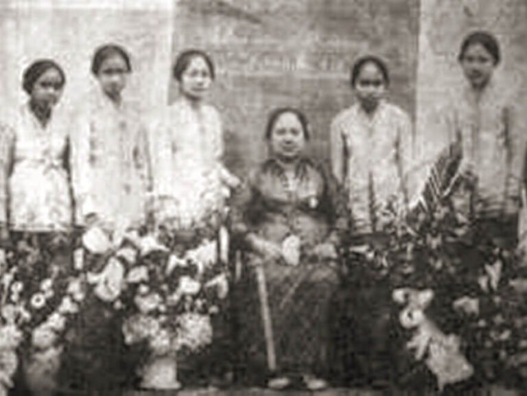 Old black-and-white group portrait: a seated elderly woman with a walking cane, surrounded by six children, with flowers and a small dog in front.