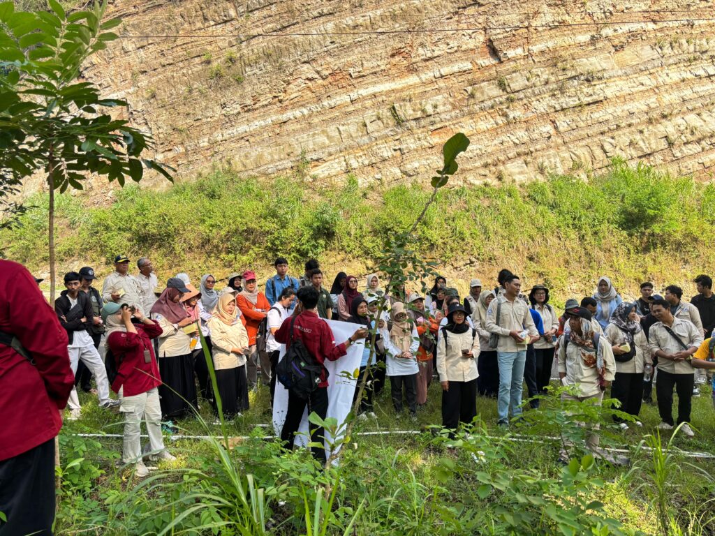 Group of students and adults gathered for an outdoor event, standing in a grassy area in front of a layered rock cliff.
