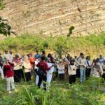 Group of students and adults gathered for an outdoor event, standing in a grassy area in front of a layered rock cliff.