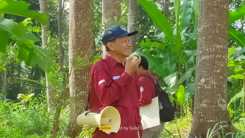 Man in a red shirt and cap speaks into a microphone in a lush tropical forest; a beige megaphone hangs from his belt.