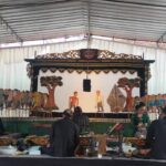 Gamelan musicians sit on the floor and play traditional Indonesian instruments in front of a carved puppetry stage backdrop.