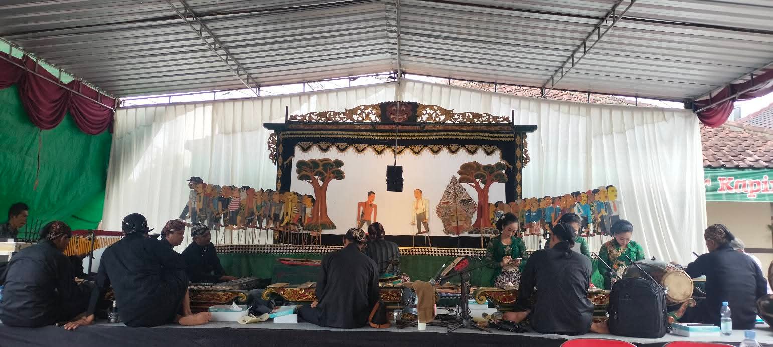 Gamelan musicians sit on the floor and play traditional Indonesian instruments in front of a carved puppetry stage backdrop.