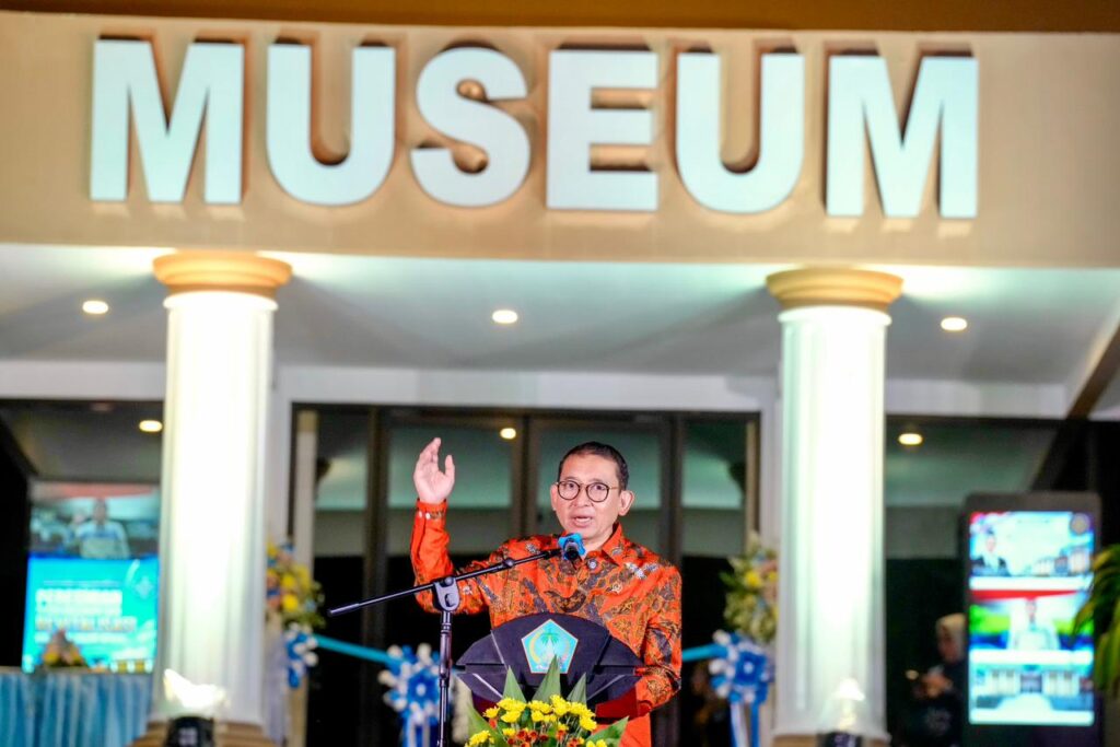 WhatsApp Image 2026-05-23 at 102013 - Mabur.co Man in red batik shirt speaks at a podium with microphone in front of a museum entrance and flowers in view.