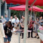 Group of people watching a man with long hair painting at an outdoor art demo under red canopies.