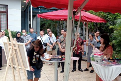 Group of people watching a man with long hair painting at an outdoor art demo under red canopies.