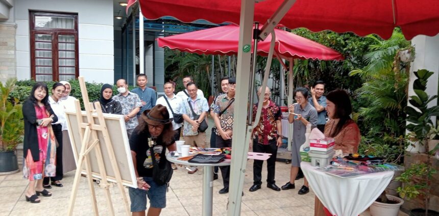 WhatsApp Image 2026-05-27 at 145450 - Mabur.co Group of people watching a man with long hair painting at an outdoor art demo under red canopies.