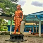 Bronze statue of a muscular ancient Indian prince standing with hands on hips on a pedestal in a plaza with blue-roofed buildings nearby.