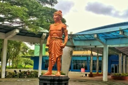 Bronze statue of a muscular ancient Indian prince standing with hands on hips on a pedestal in a plaza with blue-roofed buildings nearby.