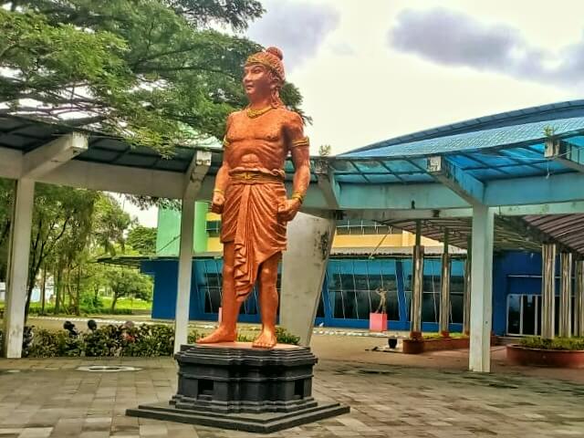 Bronze statue of a muscular ancient Indian prince standing with hands on hips on a pedestal in a plaza with blue-roofed buildings nearby.