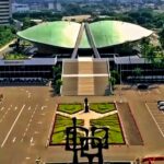 Aerial view of a large modern building with a green curved roof and a wide staircase, flanked by a landscaped plaza and statue in the foreground.