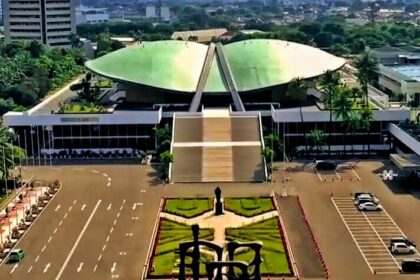 Aerial view of a large modern building with a green curved roof and a wide staircase, flanked by a landscaped plaza and statue in the foreground.