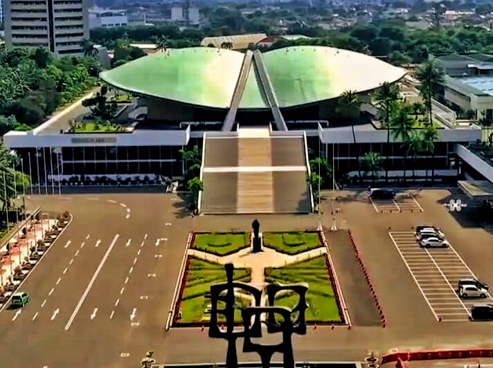 Aerial view of a large modern building with a green curved roof and a wide staircase, flanked by a landscaped plaza and statue in the foreground.