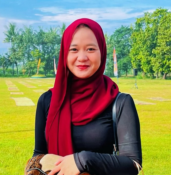 Smiling woman in a red hijab and black top standing in a sunny park with green grass and trees in the background.