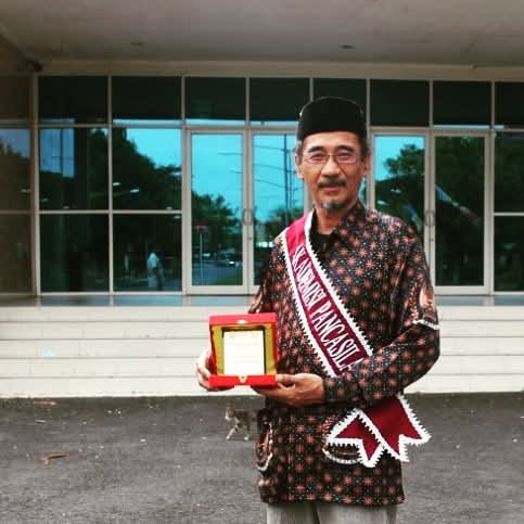 WhatsApp Image 2026-06-01 at 203311 - Mabur.co Man in a traditional batik shirt and black cap holding a red award box, wearing a sash, outside a glass-front building.